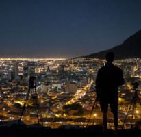 Rear View Of Silhouette Man Standing With Tripods Against Illuminated Cityscape
