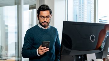 ένας άνδρας που εργάζεται στον υπολογιστή του και κοιτάζει το τηλέφωνό του