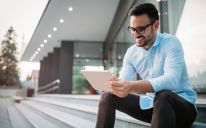 건물 앞걸이에 앉아 태블릿을 손에 들고 미소를 짓고 있는 젊은 남성 청년이 파란색 드레스 셔츠, 슬랙과 안경을 입고 있다.