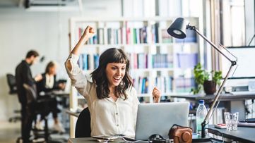 노트북을 보면서 승리의 미소를 지으며 팔을 들어 올리고 있는 여성