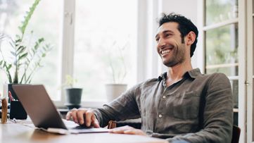 Happy businessman using laptop at table by window in home office