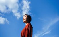 Ritratto dal basso di una giovane donna asiatica sicura di sé, in piedi sotto un bellissimo cielo azzurro limpido, che guarda verso il cielo godendosi la luce del sole. Libertà nella natura. Connessione con la natura