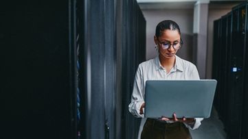 Frau steht in einem Rechenzentrum und schaut auf ihren Laptop