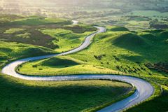 Blick von Mam Tor auf eine sich durch die hügelige Landschaft windende Straße im Abendlicht. Sommerliche grüne Wiesen und Felder.