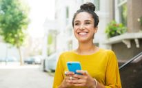 파란색 모바일 기기를 들고 미소를 지으면서 머리에 털이 있는 노란색 스웨터를 입은 젊은 여성.