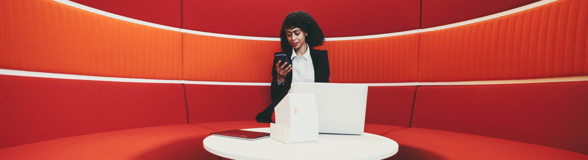 Woman sits in a bright red cubicle and works on laptop and phone.