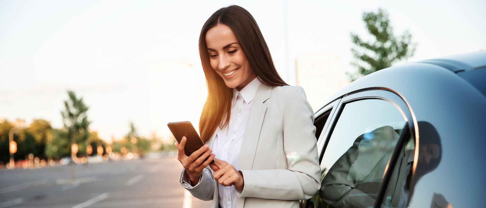 Successful smiling attractive woman in formal smart wear is using her smart phone while standing near modern car outdoors