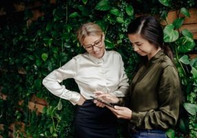 Two women looking at a tablet in front of a wall with leaves
