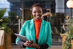 Junge, gut gekleidete Frau im offenen Lounge‑Bereich eines Bürogebäudes. Sie hält einen Laptop vor sich und lächelt direkt in die Kamera.
