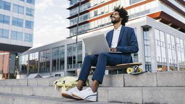 رجلٌ جالسٌ على لوحِ التزلّج الخاص به ويعمل على جهازِ الكمبيوتر الخاص به.