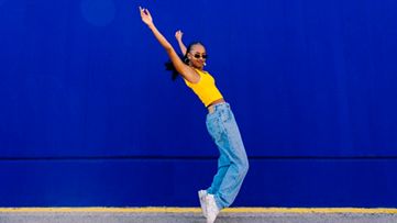 A woman dancing on her toes, wearing a yellow shirt and jeans, set against a blue background