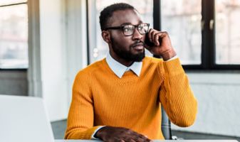 Mann im gelben Pullover sitzt vor einem Laptop und telefoniert.