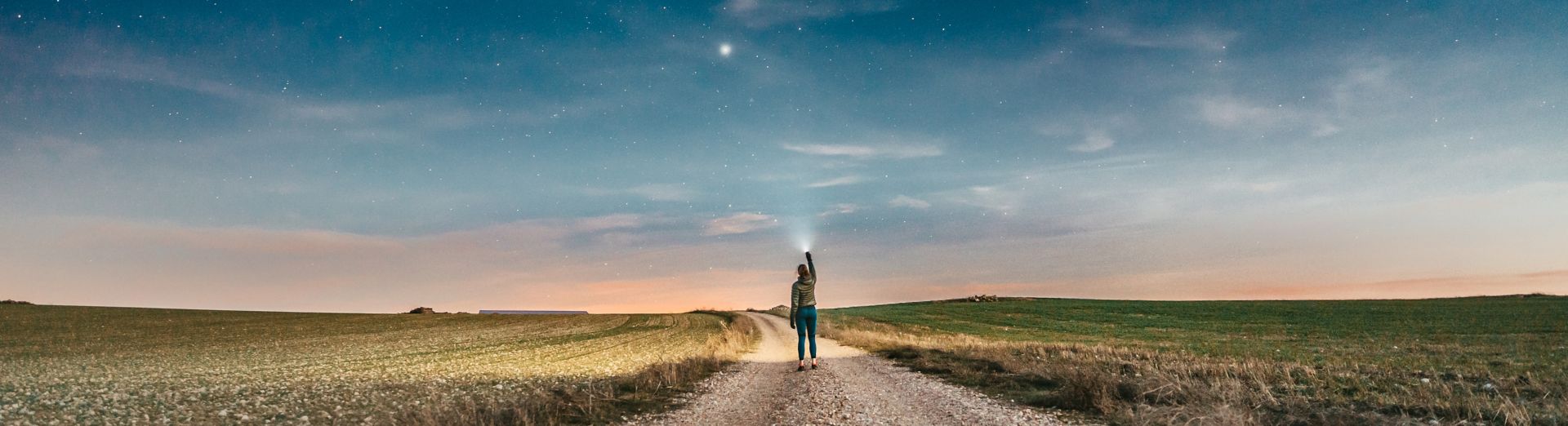 Woman walking down a dirt road at sunset, pointing at the stars with a flashlight