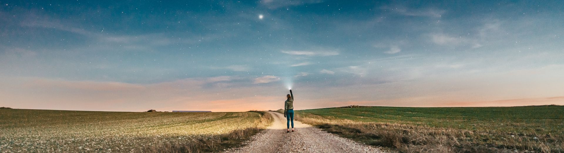 Frau geht bei Sonnenuntergang einen Feldweg entlang und zeigt mit einer Taschenlampe auf die Sterne.
